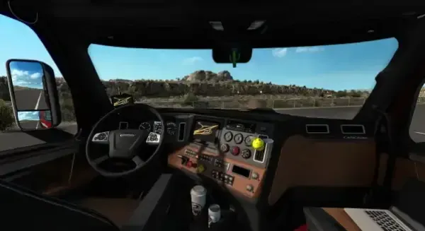 A virtual reality simulation of the interior and exterior view from inside a truck cabin, featuring a steering wheel, dashboard with various controls and displays, side mirror, and a scenic backdrop of rocky terrain under a clear blue sky. 