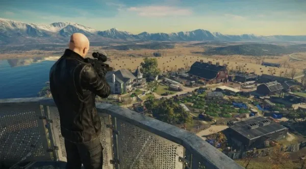 A bald man in a black leather jacket stands on a metal balcony overlooking a picturesque village nestled among rolling hills and mountains. He is holding a camera, ready to capture the serene landscape below, which includes houses with red roofs, green fields, and a body of water reflecting the clear blue sky. The scene exudes tranquility and natural beauty.