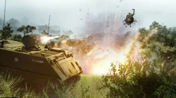 A U.S. Army tank in a grassy field firing at an enemy position, with explosions and smoke in the background, and a helicopter hovering above. The scene is set during daylight hours, with sunlight filtering through trees and casting shadows on the ground.