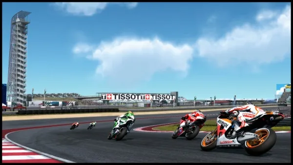 A high-speed motorcycle race on a track with riders in colorful racing suits and bikes. The scene includes a prominent Tissot advertisement, a tall tower structure, and a clear blue sky with scattered clouds. The racers are leaning into a turn, showcasing the dynamic nature of the sport. 