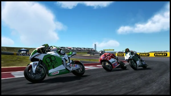 A high-speed motorcycle race on a sunny day, with three bikes in motion: one green and white, another red and white, and the third black and white. The track is lined with yellow barriers and sponsor signs like "STANLEY." Spectators are visible in the background under a clear blue sky with fluffy clouds.