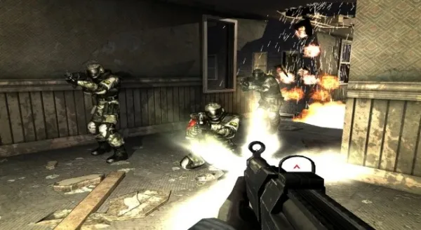 A first-person shooter video game scene featuring soldiers in combat gear engaging in a firefight inside a dilapidated building. The player's perspective shows a gun with a scope, while explosions and gunfire erupt from the background. Debris is scattered across the floor, and the environment appears war-torn with broken windows and damaged walls.
